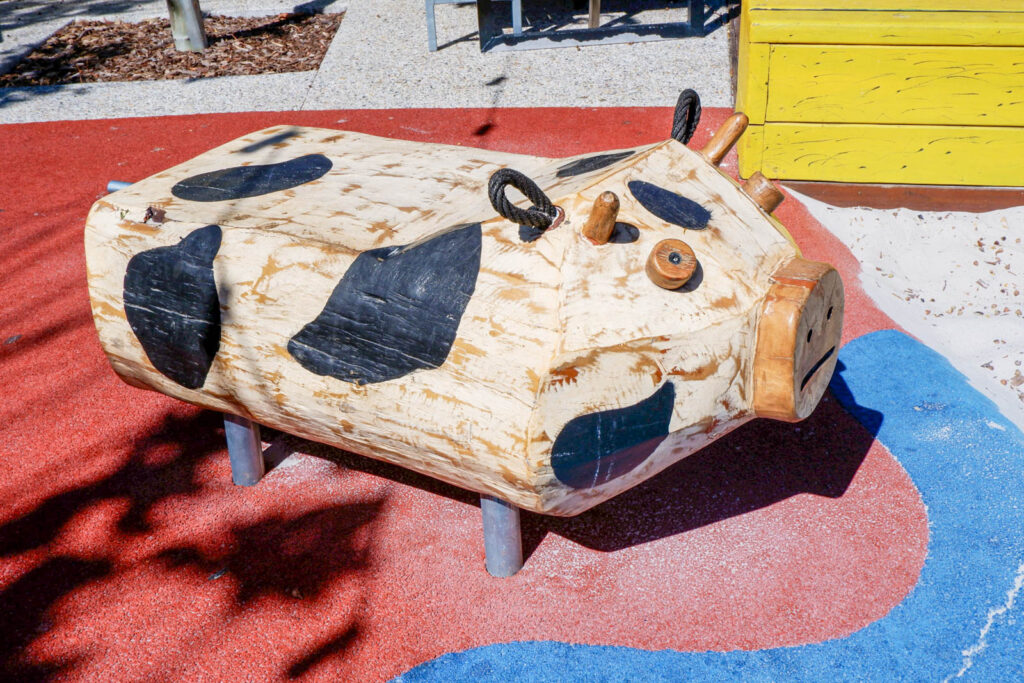 Carved Jarrah log cow with black-and-white Holstein markings on red surfacing at Charlie's Paddock playground