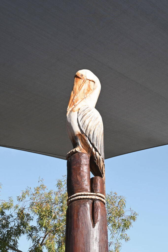 Hand-carved painted pelican sculpture at the summit of the Pelican Nest Fort, Shelley Beach Park playground — Jarrah bush pole, shade sail and blue sky