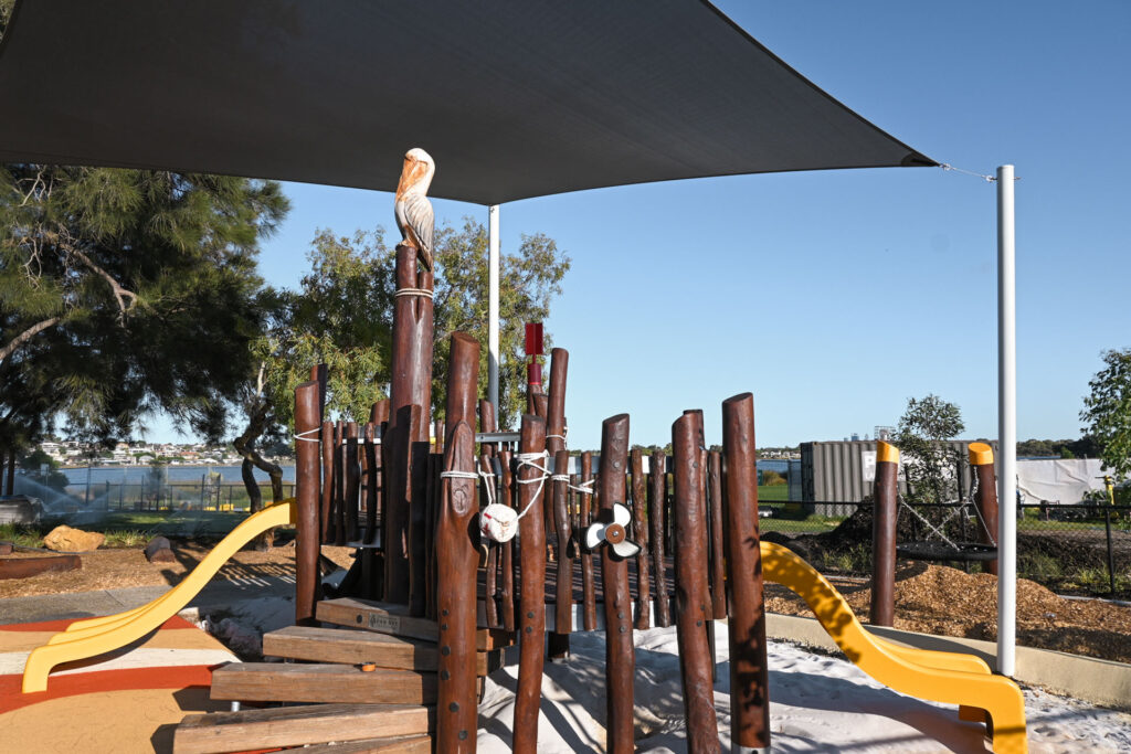 Medium-wide of the Pelican Nest Fort from ground level — pelican carving at summit of central bush pole, yellow slides either side, shade sail overhead, Canning River and far bank visible behind. Alt Text: VRI Pelican Nest Fort at Shelley Beach Park playground — carved pelican at summit of Jarrah bush pole, yellow slides and Canning River in background