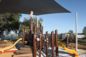Medium-wide of the Pelican Nest Fort from ground level — pelican carving at summit of central bush pole, yellow slides either side, shade sail overhead, Canning River and far bank visible behind. Alt Text: VRI Pelican Nest Fort at Shelley Beach Park playground — carved pelican at summit of Jarrah bush pole, yellow slides and Canning River in background