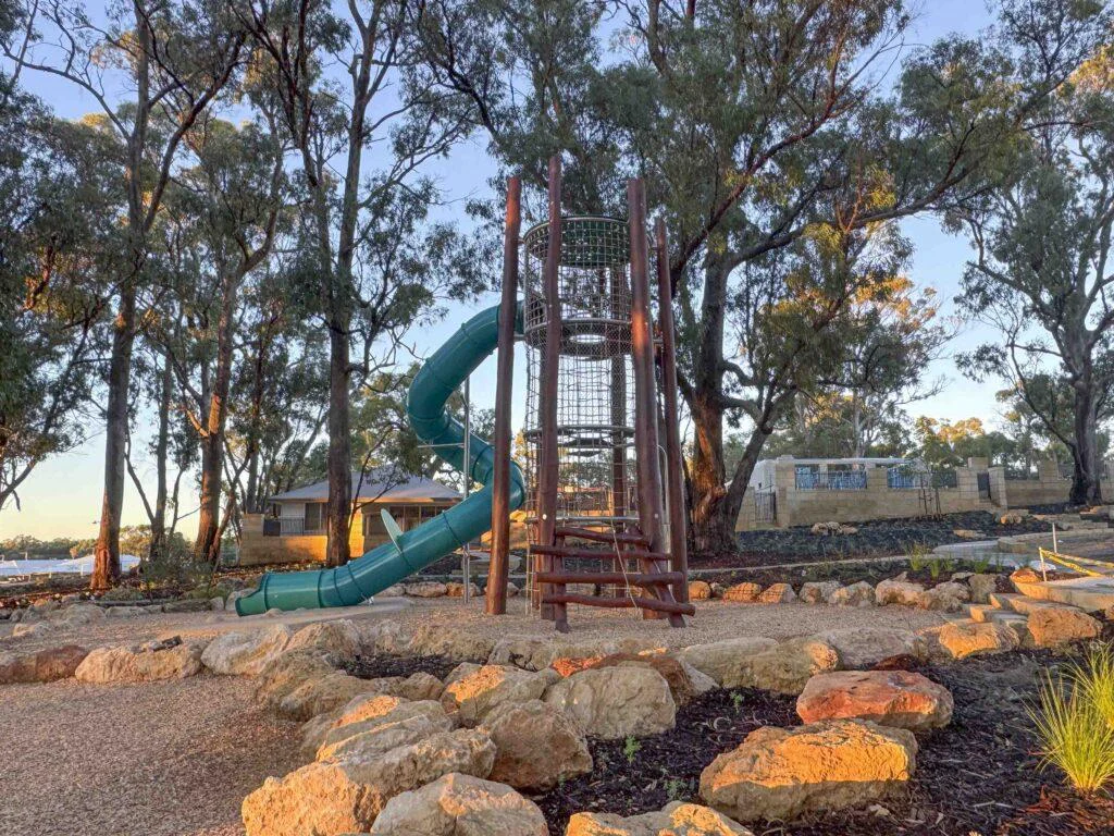 Jarrah timber climbing tower playground with teal tube slide set among mature trees and rock landscaping at Hathaway Reserve Baldivis